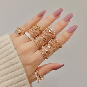 Può includere: Una mano che indossa un set di anelli d'oro con vari design, tra cui una fascia bianca, un anello a catena e un motivo a foglia. Gli anelli sono impilati su ogni dito.