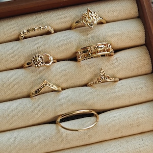 Op de afbeelding: Een verzameling goudkleurige ringen, tentoongesteld in een houten sieradendoos. De ringen hebben verschillende ontwerpen, waaronder bloemen-, ster- en geometrische patronen. De ringen zijn gerangschikt op een beige stof.