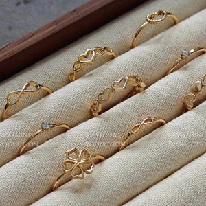 May include: A selection of gold-coloured rings presented in a wooden jewellery box. The rings showcase diverse designs, such as hearts, infinity symbols, a four-leaf clover, and a single gemstone. The rings are arranged on a cream-coloured fabric.