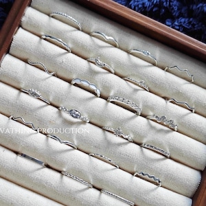 Könnte beinhalten: Eine Sammlung von Silberringen, die in einer hölzernen Schmuckschatulle ausgestellt sind. Die Ringe weisen verschiedene Designs auf, darunter Bänder mit kleinen Steinen, wellenförmige Muster und filigrane Verzierungen. Die Ringe sind auf einem cremefarbenen Stoff angeordnet.