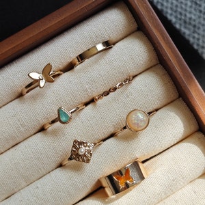 Könnte beinhalten: Eine Sammlung goldfarbener Ringe in einer Holzbox. Die Ringe weisen verschiedene Designs auf, darunter ein Schmetterling, ein Edelstein, eine Perle und ein Band mit einem orangefarbenen Schmetterling. Die Ringe liegen auf einem cremefarbenen Stoff.