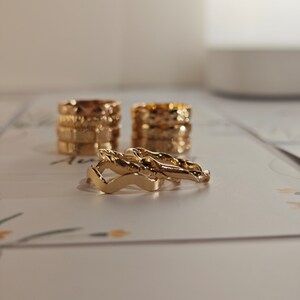 May include: A set of four gold-toned rings, stacked on top of each other. The rings are all different styles, with some featuring a textured design and others with a more simple design.