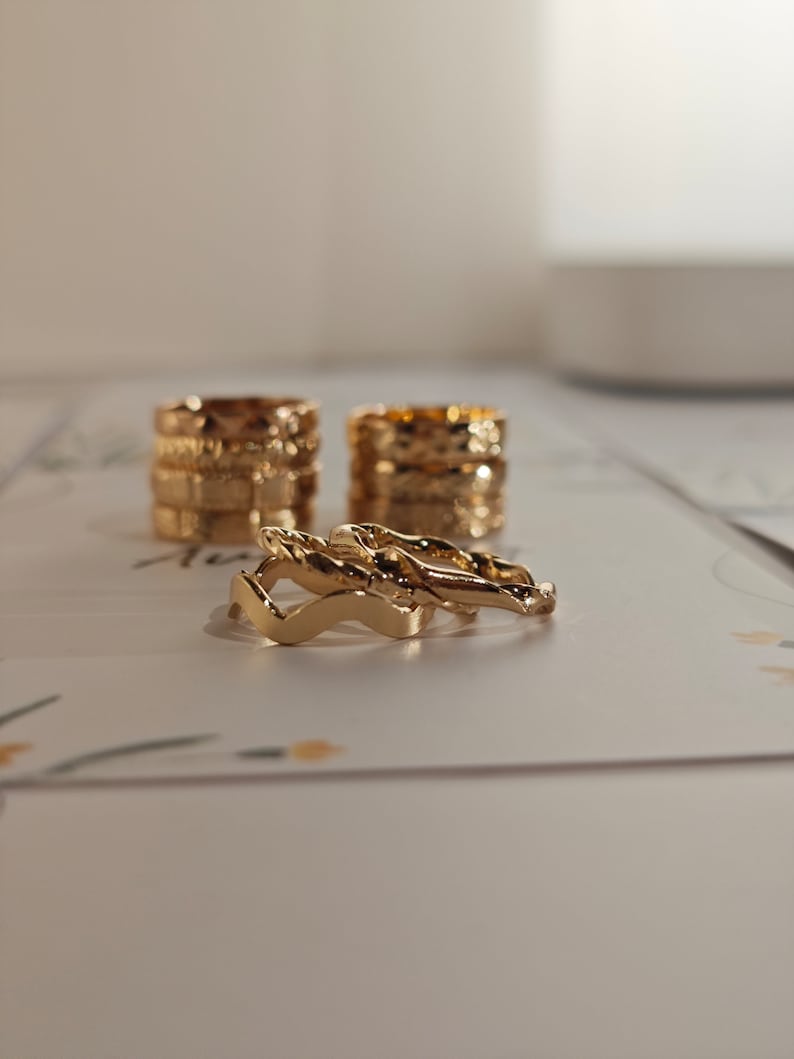 May include: A set of four gold-toned rings, stacked on top of each other. The rings are all different styles, with some featuring a textured design and others with a more simple design.