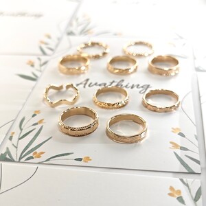 May include: A set of ten gold-toned rings with various textured designs. The rings are displayed on a white card with a floral pattern and the word "Awaiting".