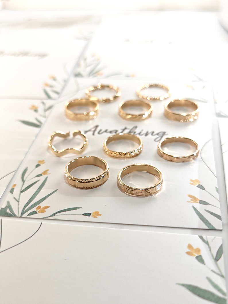 May include: A set of ten gold-toned rings with various textured designs. The rings are displayed on a white card with a floral pattern and the word "Awaiting".