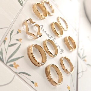 May include: A set of nine gold-toned rings with different designs, including a wavy ring, a ring with a textured surface, and a ring with a simple band. The rings are displayed on a white card with the word "Awating" printed on it.