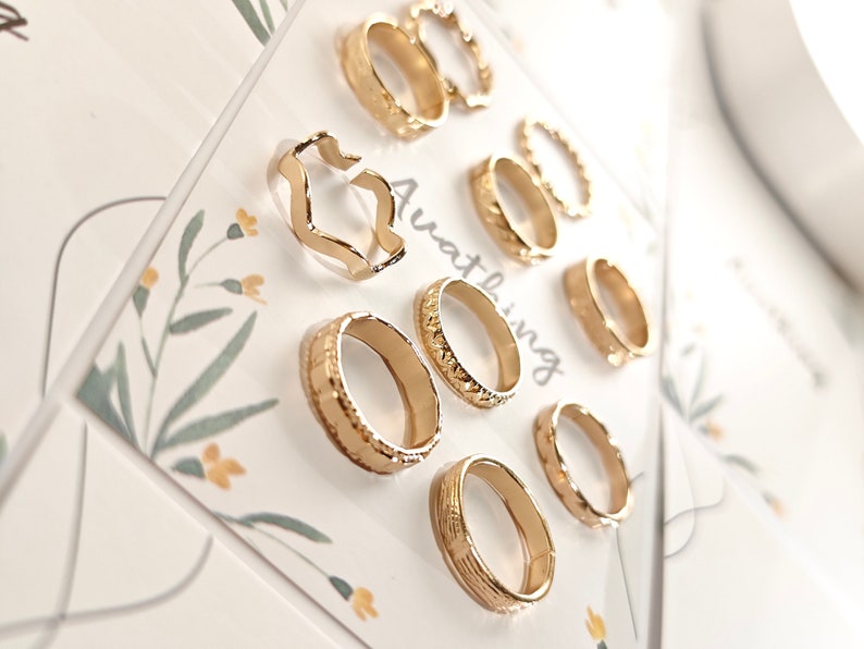 May include: A set of nine gold-toned rings with different designs, including a wavy ring, a ring with a textured surface, and a ring with a simple band. The rings are displayed on a white card with the word "Awating" printed on it.