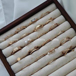 Może przedstawiać: Zestaw 20 złotych pierścionków w drewnianym pudełku z beżową podszewką z tkaniny. Pierścionki mają różne wzory, w tym serce, prostokąt, koło, falę i liść.