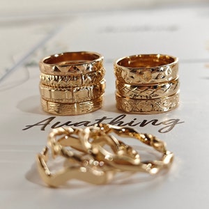 May include: A set of gold-toned rings with different textured patterns. The rings are stacked on top of each other and are arranged in a row. The rings are on a white surface with the word "Awaiting" written on it.