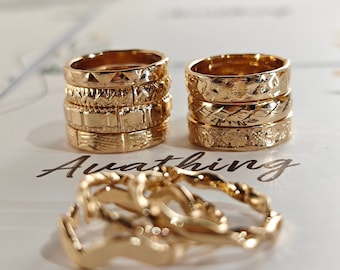 May include: A set of gold-toned rings with different textured patterns. The rings are stacked on top of each other and are arranged in a row. The rings are on a white surface with the word "Awaiting" written on it.