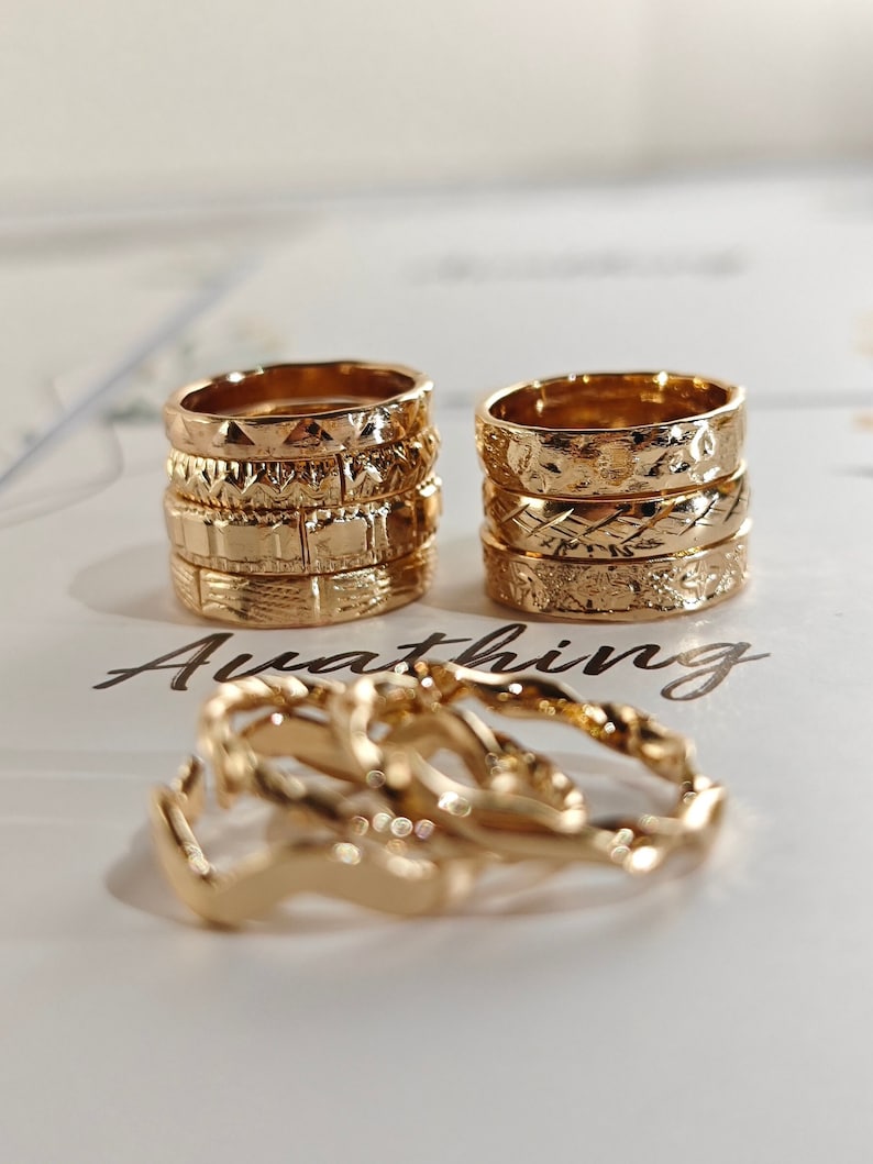 May include: A set of gold-toned rings with different textured patterns. The rings are stacked on top of each other and are arranged in a row. The rings are on a white surface with the word "Awaiting" written on it.
