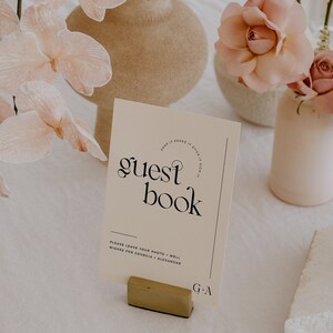 May include: A white card with black text that reads "SNAP IT SHAKE IT STICK IT SIGN IT" in a circle around the words "guest book". Below the text is "PLEASE LEAVE YOUR PHOTO + WELL WISHES FOR GEORGIA + ALEXANDAR" and "G+A". The card is sitting on a gold stand.