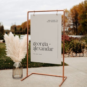 May include: A copper pipe frame holds a white sign with the text "Welcome to our wedding" and "Georgia + Alexander" with the date "11th May 20XX".