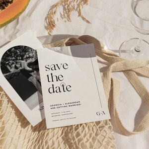 May include: Save the date wedding announcement card with a black and white photo of a couple in a modern minimalist style. The card features the text "save the date" in a bold, sans-serif font. The text "Georgia + Alexandar are getting married!" is printed below the photo. The date and location of the wedding are listed below the text. The card is printed on a white background with a black border.