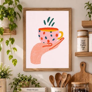 May include: A framed wall art print featuring a stylised illustration of a hand holding a pink coffee mug with blue and orange polka dots. Above the mug are green steam marks. The artwork is rendered in a textured, hand-drawn style on a light background.