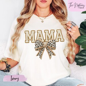 Könnte beinhalten: Elfenbeinfarbenes T-Shirt mit dem Wort "MAMA" in einem Schriftstil aus Seil und einer Schleife mit Leopardenmuster. Das Shirt hat auch den Text "The Neptunes and Co." in der oberen rechten Ecke und das Wort "Ivory" unten links.