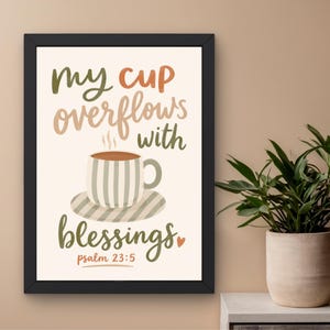May include: A framed wall art print featuring the text "my cup overflows with blessings psalm 23:5" in a mix of olive green, tan, and terracotta script fonts. A central illustration shows a striped coffee cup on a saucer with steam rising.