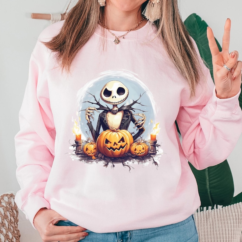 May include: A light pink sweatshirt featuring a Halloween-themed graphic. The graphic depicts Jack Skellington from The Nightmare Before Christmas, surrounded by carved pumpkins and lit candles. The design is set against a light blue background.