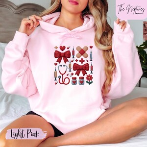 Könnte beinhalten: Hellrosa Kapuzenpullover mit medizinischem Motiv. Das Design umfasst rote Schleifen, Herzen, Pflaster, Spritzen, ein Stethoskop, Pillen und eine Blume. Der Text "Light Pink" befindet sich unten.