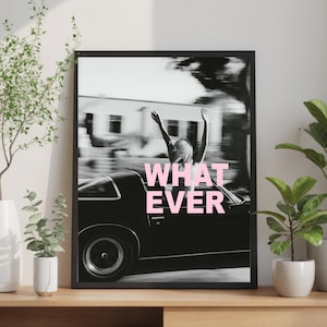 May include: A black and white framed wall art print featuring a person with arms raised in a convertible car, with the text WHAT EVER in bold pink letters centred over the image. The print is displayed in a simple black frame on a wooden surface.