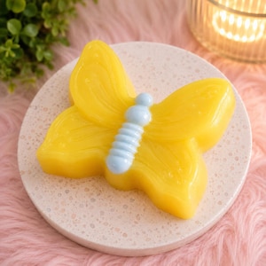 May include: A butterfly-shaped soap bar featuring bright yellow wings with subtle embossed vein details and a smooth, light blue segmented body. The soap has a glossy finish and is positioned centrally on a round, speckled off-white stone coaster.