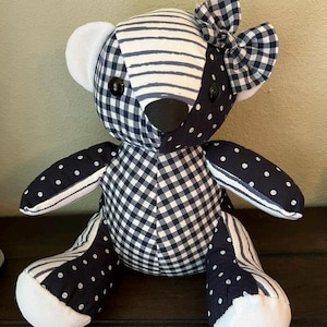 May include: A stuffed teddy bear constructed from patchwork fabric featuring navy blue and white patterns including gingham checks, polka dots, and horizontal stripes. The bear has black button eyes, a black nose, and a matching gingham bow on its ear.