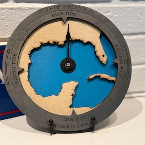 May include: A round wooden tide clock with a blue center representing the Gulf of America. The clock face is gray with text indicating high and low tide, and rising and falling tide. The clock has a black hand and is mounted on a black stand.
