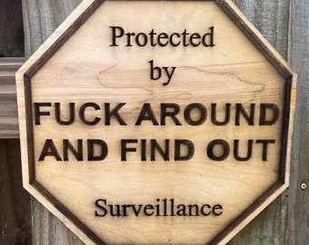 Può includere: Un cartello di legno di forma ottagonale con il testo "Protected by FUCK AROUND AND FIND OUT Surveillance". Il cartello è marrone con lettere nere.
