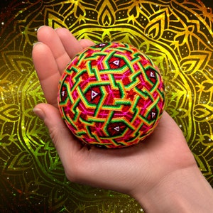 Può includere: Una mano tiene una palla rotonda, colorata, cucita a mano con un motivo geometrico. La palla è fatta di filo rosso, verde, giallo e rosa. Lo sfondo è un motivo a mandala dorato e giallo.