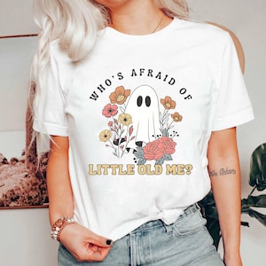 May include: White t-shirt featuring a ghost illustration surrounded by flowers. The text on the shirt reads "WHO'S AFRAID OF LITTLE OLD ME?" in a retro font. The design includes a ghost with black eyes and a floral arrangement.