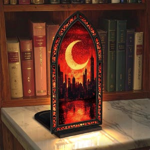 May include: A decorative stained glass bookend featuring a cityscape silhouette under a crescent moon against a red sky. The bookend has a black base and a decorative border. It is displayed on a marble surface in front of a bookshelf.