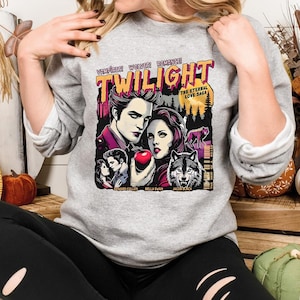 May include: A gray sweatshirt featuring a colorful graphic from the movie "Twilight." The graphic includes the title "Twilight" in large, stylized letters, along with images of the main characters, a wolf, and an apple. The text "The Eternal Love Saga" is also present.