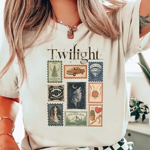 May include: Cream-colored t-shirt with the word "Twilight" in a vintage font. The shirt features a collage of postage stamp-style graphics, including a truck, wolf, and other themed illustrations. The design is printed on a soft, light-colored fabric.