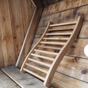 May include: A light-colored wooden sauna headrest with a curved design and multiple horizontal bars. The headrest is leaning against a wooden wall, showcasing the natural wood grain and texture. The number "8" is engraved on the bottom.