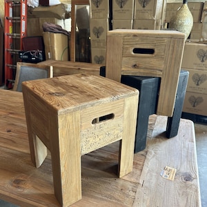 May include: Two square wooden stools with a natural light brown finish and visible wood grain. Each stool features a rectangular cutout handle on the side panel. One stool is positioned in the foreground, while the second sits atop a black base.
