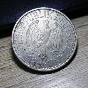 Op de afbeelding: Een zilveren munt met een adelaar en de tekst "DEUTSCHE BUNDESREPUBLIK". De munt is licht versleten, maar de details van het ontwerp zijn zichtbaar. De munt ligt op een houten oppervlak.