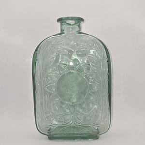 May include: A clear, light green glass bottle with a flattened, rectangular shape and a narrow neck. The front surface features a molded, embossed design of a large central sunflower surrounded by detailed leaves and stems in relief.