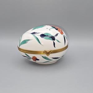 May include: An egg-shaped, hinged trinket box with a white base, adorned with floral and leaf designs in teal, navy, and orange. The box features a gold-colored band around the middle and a decorative clasp.