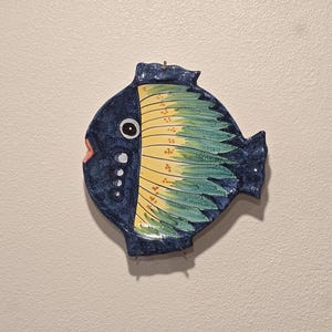 May include: A ceramic fish wall hanging. The fish is primarily dark blue with yellow and green accents. The fish has a large eye, a small pink mouth, and a decorative pattern on its body. A small wire loop is at the top for hanging.
