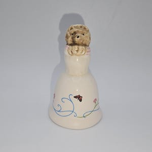 May include: A ceramic bell with a bear figurine on top. The bell is cream-colored with blue swirls, pink flowers, and a butterfly design. The bear is light brown with textured fur.