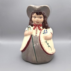 May include: A ceramic figurine of a girl wearing a hat and a dress. The figurine is painted with a cream-colored dress with red trim, a gray skirt, and a gray hat. She holds a shepherd's crook and a sprig of blue flowers.