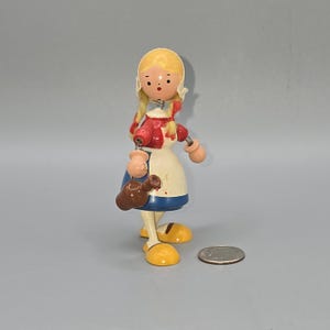 May include: A vintage wooden doll with a blonde hairstyle, wearing a red jacket, white apron, and blue skirt. The doll is holding a brown jug and has yellow shoes. The doll is standing on a gray surface.