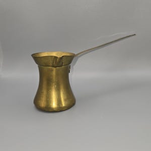 May include: A vintage brass coffee pot with a long, slender handle. The pot has a rounded base that tapers to a narrow waist, then flares out to a wide, fluted rim. The brass has a warm, golden hue, suggesting age and use. The handle extends horizontally.