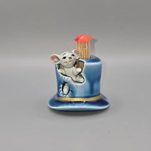 May include: A whimsical, vintage toothpick holder featuring a blue top hat design with a gold band. A small, gray mouse figurine peeks out from the hat. A small red umbrella tops the toothpicks. A charming collectible.