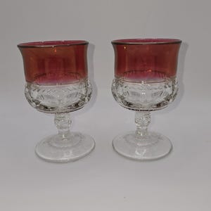 May include: Two clear glass wine goblets with a burgundy red rim. The goblets have a cut glass design and a short stem.