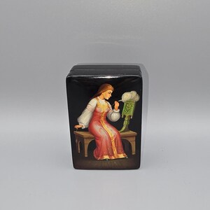 May include: A black wooden box with a painted scene of a woman in a red dress sitting on a bench. The woman is looking at a green object with a white cloud above it.