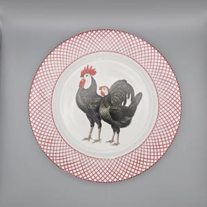 May include: A white ceramic plate with a red and white lattice border. The center of the plate features a detailed illustration of two roosters in shades of black, gray, and red. A decorative kitchenware item.
