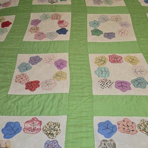 May include: A handmade quilt with a green background and white squares. Each square features a circle of appliqued leaf shapes in various colors and patterns, including red, blue, and floral prints. The quilt has a traditional, handcrafted aesthetic.