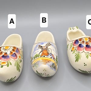May include: Three ceramic Dutch clogs, each with a unique hand-painted design. The clogs are off-white with floral and windmill motifs in various colors. The clogs are labeled A, B, and C.