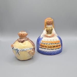 May include: Two ceramic figurines. One is a round figure in a yellow robe with a brown shawl and a basket. The other is a tall figure in a blue dress with orange and black accents, a white apron, and blonde hair.